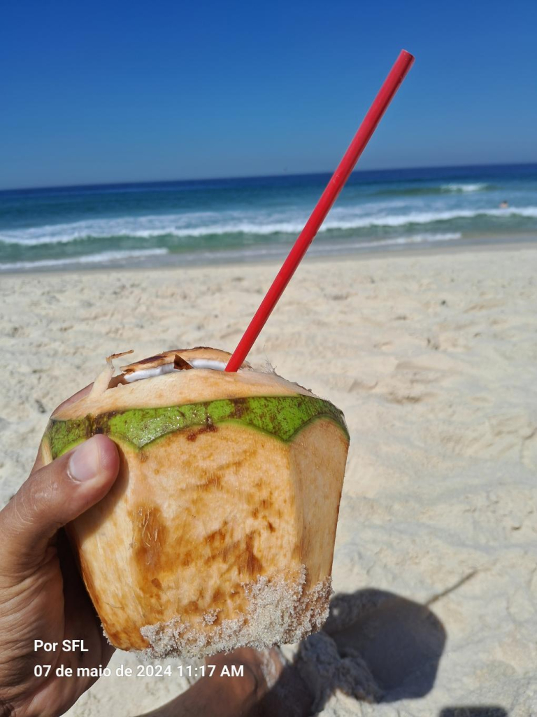 foto de um coco descascado com um canudo vermelho tendo ao fundo as areias da praia, uma amorinha e o céu azul de um dia de sol. Ele é segurado por uma mão humana.